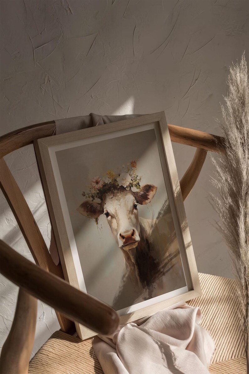 May include: A framed painting of a white cow wearing a flower crown. The cow is looking directly at the viewer with a gentle expression. The background is a light brown colour.