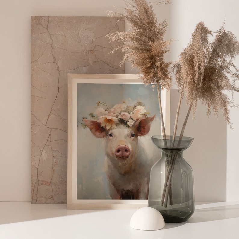 Puede incluir: Una pintura enmarcada de un cerdo con una corona de flores. El cerdo es blanco con orejas rosadas y un hocico rosado. El fondo es azul claro con una nube blanca. La pintura est&aacute; en un marco blanco y est&aacute; sobre una superficie blanca. Hay un jarr&oacute;n con hierba de la pampa seca en el fondo.