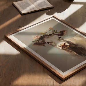 May include: A framed print of a cow wearing a flower crown. The cow is white with brown patches and has a gentle expression. The background is a soft, muted grey.