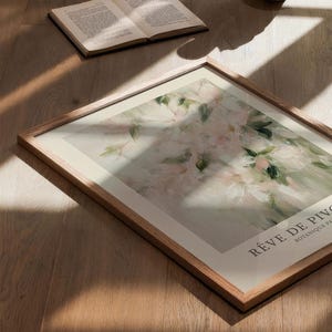 May include: A framed floral art print with a wooden frame, featuring a soft, impressionistic painting of pink and white flowers with green leaves. The text "R&Ecirc;VE DE PIVO" is visible. An open book and a white vase are also present.