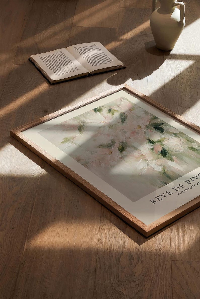 May include: A framed floral art print with a wooden frame, featuring a soft, impressionistic painting of pink and white flowers with green leaves. The text "R&Ecirc;VE DE PIVO" is visible. An open book and a white vase are also present.