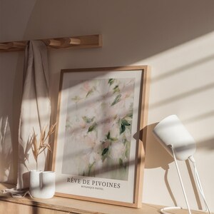May include: A framed art print featuring a soft, abstract floral design in shades of pink and green. The print is displayed on a wooden cabinet, with a white lamp and a small vase with dried plants. The text on the print reads "R&ecirc;ve de Pivoines".