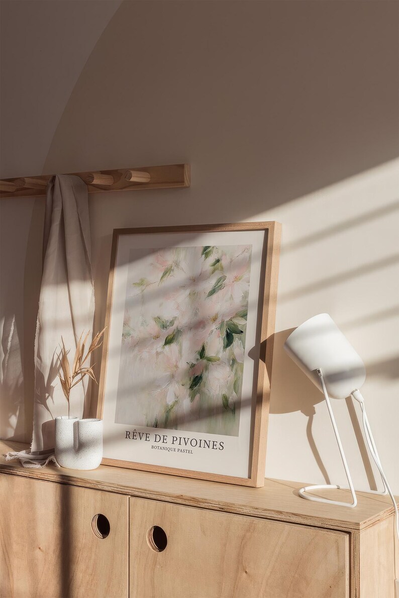 May include: A framed art print featuring a soft, abstract floral design in shades of pink and green. The print is displayed on a wooden cabinet, with a white lamp and a small vase with dried plants. The text on the print reads "R&ecirc;ve de Pivoines".