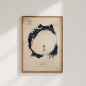 Puede incluir: Obra de arte enmarcada con una ilustración de rana en tinta negra. La rana está representada en un estilo minimalista con un cuerpo circular y líneas simples. También hay texto japonés y sellos rojos.