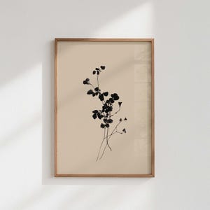 Könnte beinhalten: Ein gerahmter botanischer Druck mit minimalistischem Design. Das Kunstwerk zeigt eine zarte schwarze Blumenanordnung vor einem neutralen beigefarbenen Hintergrund. Der Rahmen besteht aus hellbraunem Holz und verleiht dem Stück Wärme.