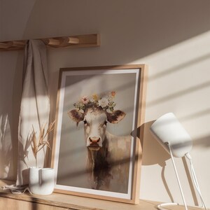 May include: A framed print of a white cow wearing a flower crown. The cow is looking at the camera. The print is on a wooden shelf with a white lamp beside it.