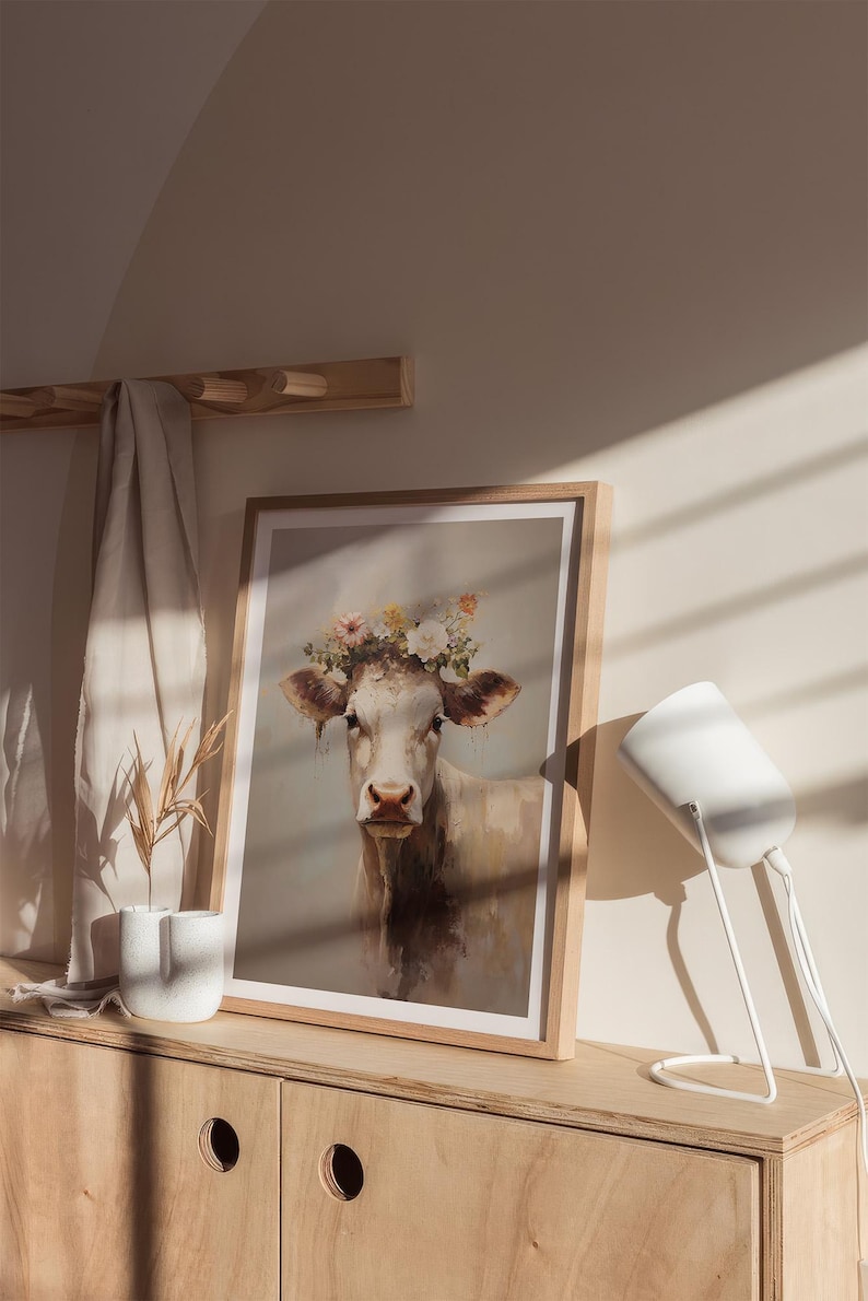 May include: A framed print of a white cow wearing a flower crown. The cow is looking at the camera. The print is on a wooden shelf with a white lamp beside it.