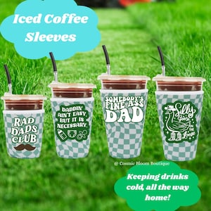 May include: Four iced coffee cups with black straws and decorative sleeves. The sleeves have various designs and text, including "RAD DADS CLUB," "DADDIN AIN'T EASY," "SOMEBODY'S FINE ASS DAD," and "Silly Goose on the Loose." The background is green grass.