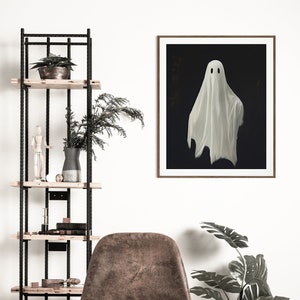 Peut inclure: Une impression encadr&eacute;e d'un fant&ocirc;me blanc avec deux yeux noirs sur un fond sombre. L'impression est accroch&eacute;e &agrave; un mur blanc au-dessus d'un fauteuil en cuir marron et d'une plante en pot.