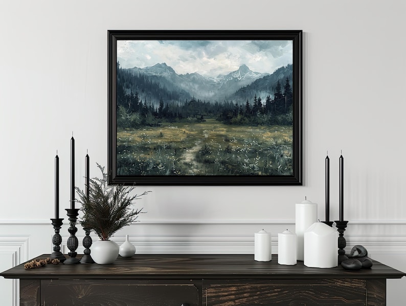 K&ouml;nnte beinhalten: Ein gerahmtes Landschaftsbild mit einer Bergkette und einem Wald im Vordergrund. Das Bild ist in einem schwarzen Rahmen und h&auml;ngt an einer wei&szlig;en Wand. Vor dem Bild stehen Kerzen und eine Pflanze auf einem dunklen Holztisch.
