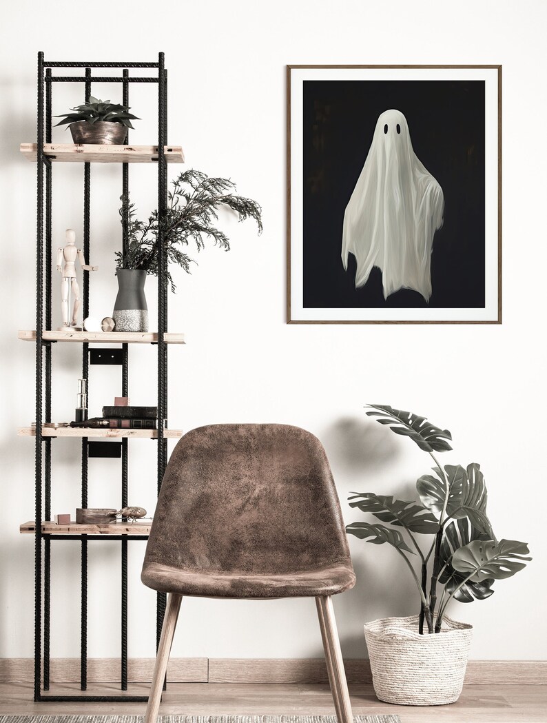 Peut inclure: Une impression encadr&eacute;e d'un fant&ocirc;me blanc avec deux yeux noirs sur un fond sombre. L'impression est accroch&eacute;e &agrave; un mur blanc au-dessus d'un fauteuil en cuir marron et d'une plante en pot.