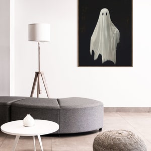 Peut inclure: Un fant&ocirc;me blanc avec deux yeux noirs est peint sur une toile noire. La toile est encadr&eacute;e dans un cadre en bois fonc&eacute; et est accroch&eacute;e &agrave; un mur blanc.