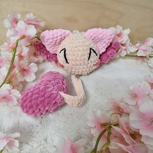 Peut inclure: Une peluche axolotl crochetée à la main, aux tons rose et beige, avec des détails brodés en noir. La peluche est entourée de fleurs de cerisier roses sur une surface blanche et duveteuse. L'axolotl a une tête ronde, de grandes oreilles et un visage souriant.