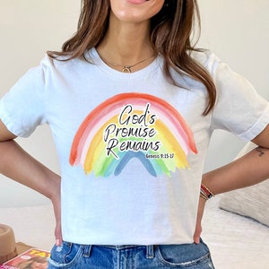 Pu&ograve; includere: Una maglietta bianca con un design arcobaleno e il testo "God's Promise Remains" con il riferimento biblico "Genesi 9:15-17".