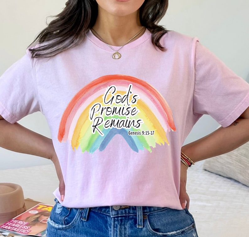 Pu&ograve; includere: Maglietta rosa con un design arcobaleno e il testo "God's Promise Remains" e "Genesis 9:15-17".