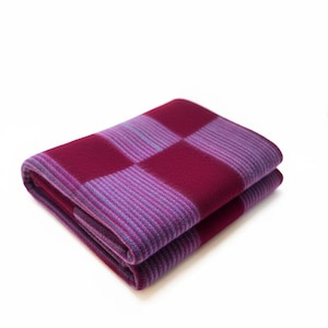 Può includere: Coperta piegata con un motivo a scacchiera nei toni del bordeaux e del viola. La coperta è realizzata in un materiale morbido ed è piegata con cura, mettendo in risalto le variazioni di colore e la trama. Lo sfondo è bianco.