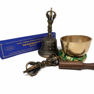May include: A selection of meditation items: a brass singing bowl, a dark metal bell, a wooden mallet, a dorje, and a blue box of incense with the word "Tranquility". The bowl sits on a green cushion.