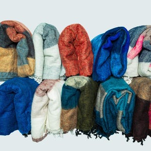 Op de afbeelding: Een verzameling opgevouwen sjaals in verschillende kleuren, waaronder blauw, oranje en grijs. De sjaals lijken gemaakt te zijn van een zacht, getextureerd materiaal en zijn in een stapel gerangschikt. De afbeelding is goed belicht en de kleuren zijn levendig.