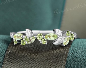 May include: A silver ring with a leaf and vine pattern, embellished with light green gemstones. The ring showcases small leaves and marquise-cut stones. The background is teal and green.