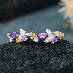 May include: A silver ring featuring a leaf design with purple and yellow gemstones. The ring has a delicate, nature-inspired aesthetic, with the gemstones set among the leaves. The ring is displayed on a dark green surface.
