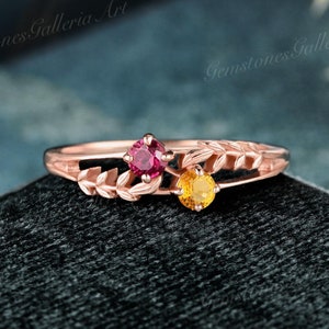 May include: A delicate rose gold ring with a leafy design. The ring features two small gemstones, one red and one yellow.