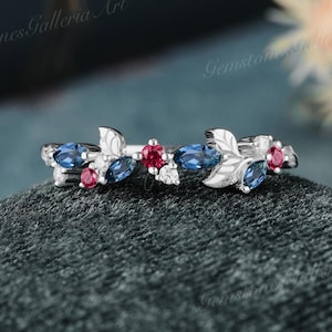 May include: A delicate silver ring with a floral design featuring blue and red gemstones. The ring is set on a gray velvet background.