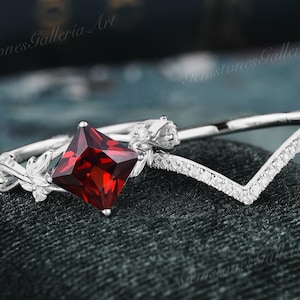 May include: A silver ring set featuring a princess-cut, deep red gemstone and a V-shaped band with small, clear stones. The main ring has a floral design on the side. The rings are displayed on a dark green velvet surface.