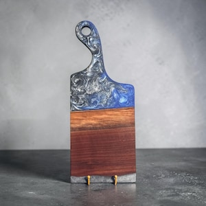 May include: A rectangular serving board featuring a dark brown wood base and a top section made of blue and silver swirling resin. The board includes a handle with a circular cutout at the top and a small gray resin accent at the bottom edge.