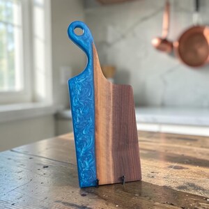 May include: A serving board featuring a combination of dark brown wood and a vibrant blue resin section with a swirling pattern. The board has a handle with a circular cutout at the top and a straight, rectangular body with a flat base.