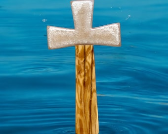 May include: A wooden cross with a silver top, standing upright in blue water. The cross is made of light brown wood with dark brown streaks.