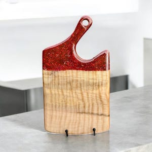 May include: A wooden cutting board with a red glitter epoxy resin top. The board has a unique shape with a handle and is displayed on a black stand.