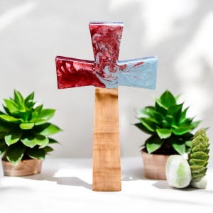 Puede incluir: Una cruz de madera con una parte superior de resina marmoleada roja y azul. La cruz está de pie sobre una superficie blanca con dos pequeñas plantas en macetas en el fondo.