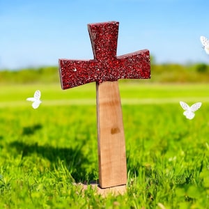 Op de afbeelding: Een rood houten kruis met glitteraccenten staat in een veld met groen gras. Witte vlinders vliegen rond het kruis.