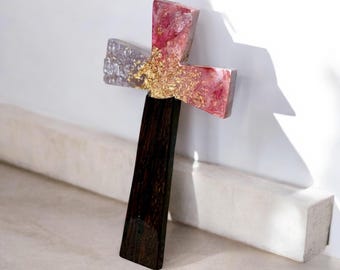 May include: A decorative cross with a dark wooden base and a crossbar made of resin. The crossbar features a mix of pink, silver, and gold leaf accents. The cross is leaning against a white wall.