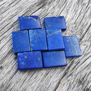 Puede incluir: Una colección de ocho pequeñas piedras rectangulares de lapislázuli de color azul con motas doradas. Las piedras están dispuestas en un patrón escalonado sobre una superficie de madera marrón claro.