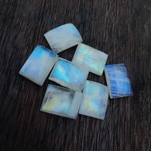 Könnte beinhalten: Eine Gruppe von sieben regenbogenfarbenen Mondstein-Cabochons, geschnitten in rechteckige Formen, mit einem Farbschema aus Weiß, Blau und Gelb. Die Steine sind in einer Gruppe auf einer dunklen Holzoberfläche angeordnet.
