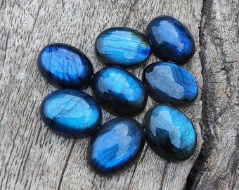 May include: A group of eight oval-shaped labradorite gemstones in shades of blue and green. The stones are arranged in a cluster on a wooden surface.