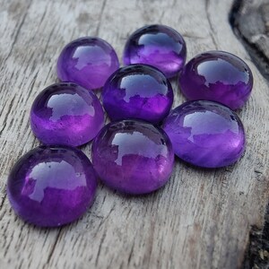 May include: Eight round, polished amethyst gemstones in various shades of purple, arranged in a cluster on a weathered wooden surface.