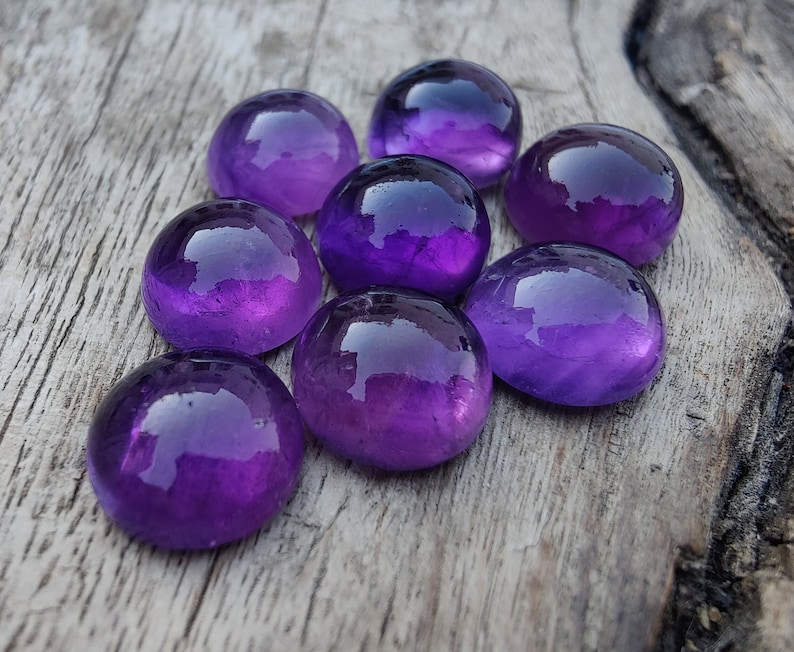 May include: Eight round, polished amethyst gemstones in various shades of purple, arranged in a cluster on a weathered wooden surface.