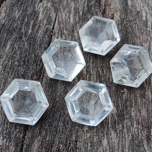 May include: Five clear, faceted, hexagonal gemstones on a gray wooden surface.