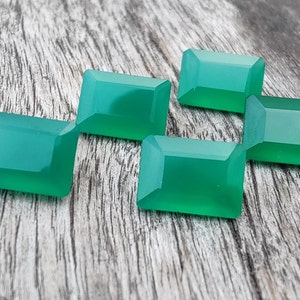 May include: Five emerald green gemstone cabochons, cut in a rectangular shape, are arranged on a wooden surface.