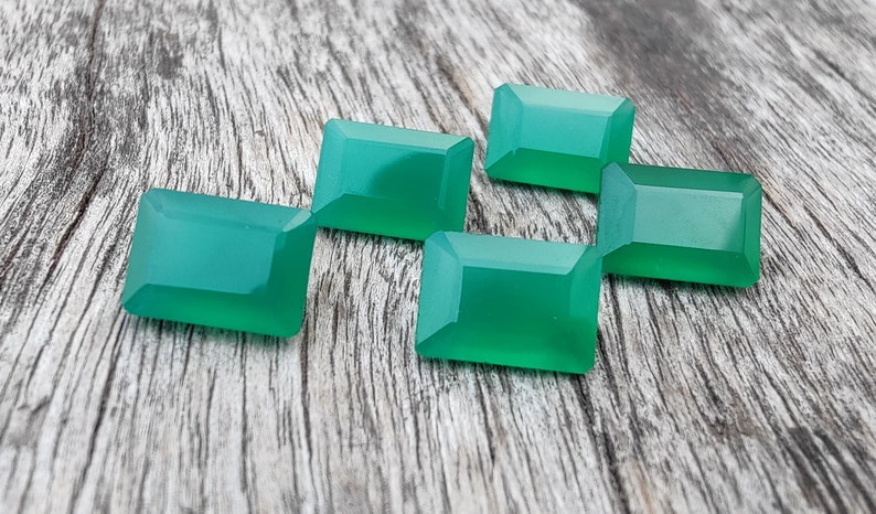 May include: Five emerald green gemstone cabochons, cut in a rectangular shape, are arranged on a wooden surface.