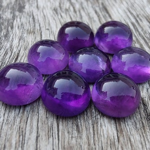 May include: Eight round, polished amethyst stones in various shades of purple, arranged in a cluster on a weathered wooden surface.