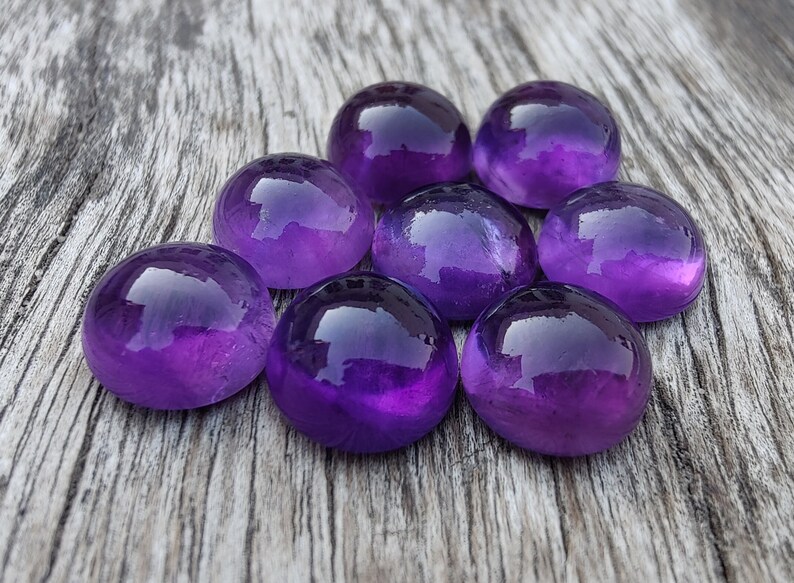 May include: Eight round, polished amethyst stones in various shades of purple, arranged in a cluster on a weathered wooden surface.
