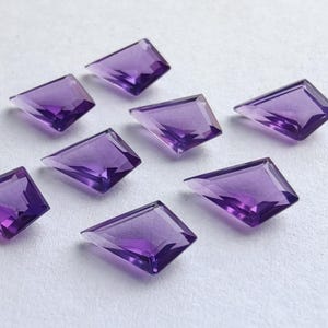 Puede incluir: Ocho gemas de amatista facetadas con forma de cometa dispuestas sobre una superficie blanca. Las gemas son de color púrpura intenso y están talladas con la parte superior plana y los lados angulados.