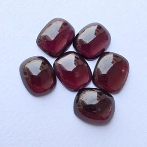 Peut inclure: Six pierres précieuses polies, de forme ovale, rouge foncé. Les pierres ont une surface lisse et brillante et reflètent la lumière. Les pierres précieuses sont disposées sur une surface blanche. Elles pourraient être utilisées pour la joaillerie ou comme éléments décoratifs.