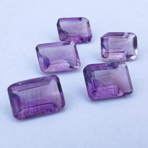May include: Five purple amethyst gemstones cut in a rectangular emerald shape. The gemstones are faceted and have a translucent appearance.
