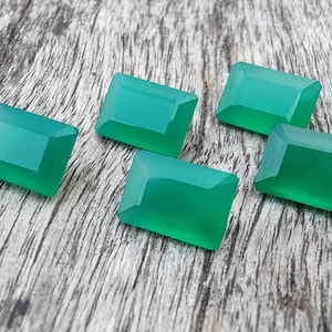 May include: Five emerald green gemstone cabochons, cut in a rectangular shape, are arranged on a wooden surface.