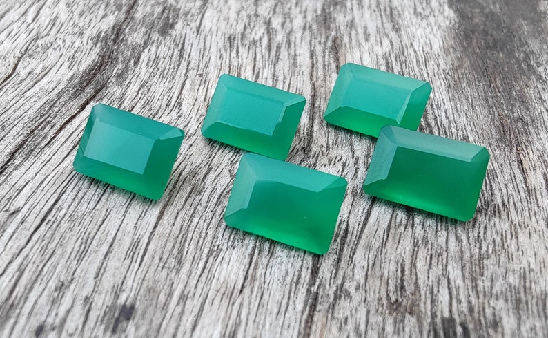 May include: Five emerald green gemstone cabochons, cut in a rectangular shape, are arranged on a wooden surface.