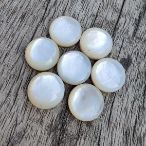 Könnte beinhalten: Acht weiße, irisierende, runde Perlmutt-Cabochons, jeder mit einem leicht unterschiedlichen Schimmer, in einem Kreis angeordnet auf einer Holzoberfläche.
